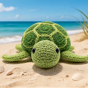 Puede incluir: Una tortuga verde de ganchillo hecha a mano, con un caparazón texturizado y ojos negros, descansa en una playa de arena. La tortuga está rodeada de conchas y arena, con el océano y el cielo azul de fondo.