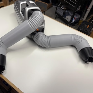 May include: A silver and black portable air filtration unit featuring two flexible, corrugated grey hoses extending from the main housing. Each hose terminates in a black, flared plastic nozzle designed for targeted air intake or exhaust.