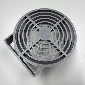 May include: A gray, circular fan grill with a square mounting bracket. The grill features concentric rings and intersecting bars, creating a grid pattern. The object is made of a solid, matte material.