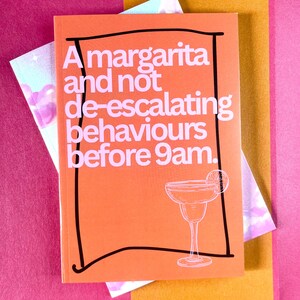 Puede incluir: Un cuaderno naranja con el texto "A margarita and not de-escalating behaviours before 9am" en blanco. Una línea negra bordea el texto y una ilustración de copa de margarita. El cuaderno está sobre un fondo rosa y dorado.