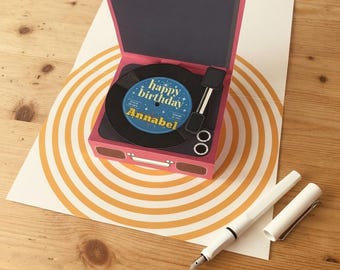May include: A pink pop-up greeting card shaped like a portable record player. The center of the black vinyl record features a blue label with the text "happy birthday" and "Annabel" in yellow and white lettering. A white fountain pen lies nearby.