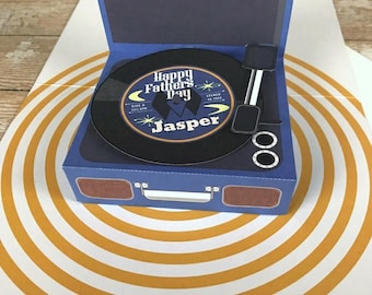 May include: A blue pop-up greeting card shaped like a vintage record player. The center label on the black vinyl record features the text Happy Fathers Day and the name Jasper. The card base includes a silver handle and two brown speakers.