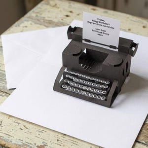 May include: A dark brown paper pop-up greeting card shaped like a vintage typewriter with a white paper insert. The insert features the printed text: To Dad, Happy Birthday!! Hope you have a great day. Love from David and Nancy xxxx. A white envelope is included.