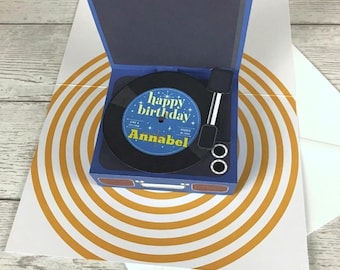 May include: A pop-up greeting card featuring a blue record player design. The black vinyl record in the center displays the text "happy birthday" and the name "Annabel" in yellow. The card base features a yellow and white concentric circle pattern.