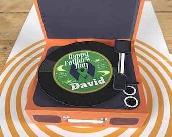May include: A pop-up greeting card shaped like a vintage record player with an orange exterior. The black vinyl record features a green center label with the text Happy Fathers Day and David, along with a black tonearm and two white knobs.