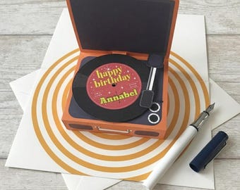 May include: A three-dimensional pop-up greeting card shaped like a vintage record player in orange and dark gray. The circular record label is red with the text "happy birthday" and "Annabel" in yellow. A white fountain pen lies nearby.