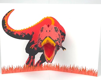 May include: A pop-up greeting card featuring a red and black Tyrannosaurus Rex dinosaur with an open mouth, yellow teeth, and orange accents. The card is set against a white background with orange grass.