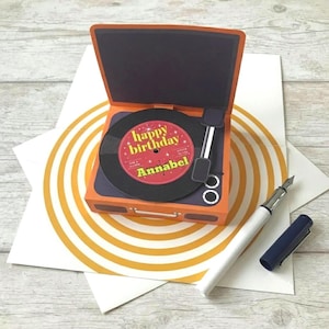 May include: A three-dimensional pop-up greeting card shaped like a vintage record player in orange and dark grey. The circular record label is red with yellow text reading happy birthday Annabel. A white fountain pen with a dark blue cap lies nearby.