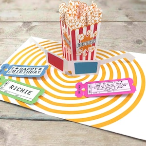 May include: A pop-up greeting card featuring a red and white striped popcorn bucket, 3D glasses with red and blue lenses, and three ticket-shaped cutouts. The tickets are blue, green, and pink, with text reading Happy Birthday, Richie.