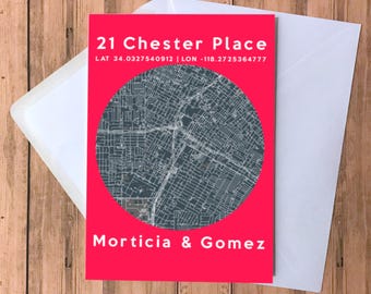 May include: A greeting card with a red background featuring a map design inside a circle. The card includes the address "21 Chester Place" and the names "Morticia & Gomez". The card is inside a white envelope, set against a wooden background.