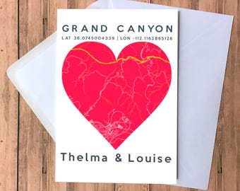 May include: A white card with a red heart-shaped map of Grand Canyon National Park. The text "GRAND CANYON" is at the top of the card, and the coordinates "LAT 36.0745004339 | LON -112.1162865126" are below. The text "Thelma & Louise" is at the bottom of the card.