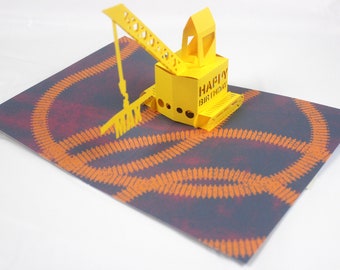 May include: A yellow pop-up birthday card featuring a construction crane. The card has the words "HAPPY BIRTHDAY" on the crane. The background is a dark gray with an orange pattern.