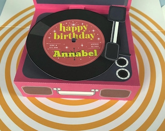 May include: A pink paper record player gift box featuring a black vinyl disc with a red center label. The label displays the text happy birthday and the name Annabel in yellow lettering, along with star accents, a tone arm, and speaker details.