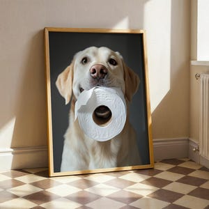 May include: A framed print featuring a golden Labrador Retriever holding a roll of white toilet paper in its mouth. The dog has a light brown coat and is set against a dark gray background. The frame is a light brown color.