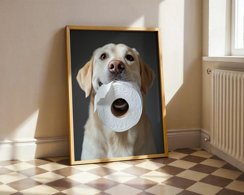 May include: A framed print featuring a golden Labrador Retriever holding a roll of white toilet paper in its mouth. The dog has a light brown coat and is set against a dark gray background. The frame is a light brown color.