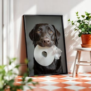 Puede incluir: Impresi&oacute;n enmarcada de un Labrador Retriever color chocolate sosteniendo un rollo de papel higi&eacute;nico en la boca. El perro tiene ojos marrones y un marco negro. La impresi&oacute;n est&aacute; apoyada contra una pared blanca junto a un peque&ntilde;o taburete blanco con una planta en maceta.