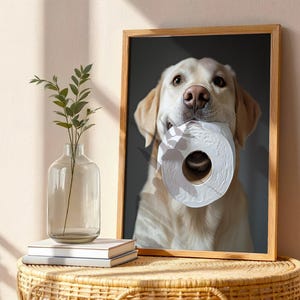 May include: A framed print featuring a golden Labrador Retriever holding a roll of white toilet paper in its mouth. The dog's expression is focused, with a light background and a wooden frame.
