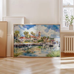 May include: Watercolor painting of a coastal town with a harbor, boats, and palm trees. The artwork features a light-colored building with a clock tower, and the water reflects the sky and buildings. The painting is in a wooden frame.
