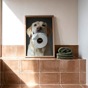 May include: A framed print featuring a golden Labrador Retriever holding a roll of toilet paper in its mouth. The dog is centered in the frame, with a neutral background. The print is displayed on a tiled surface.