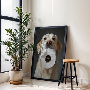 May include: A framed print featuring a golden retriever holding a roll of toilet paper in its mouth. The dog is centered in the frame against a dark background. The print is displayed next to a potted plant and a small wooden stool.