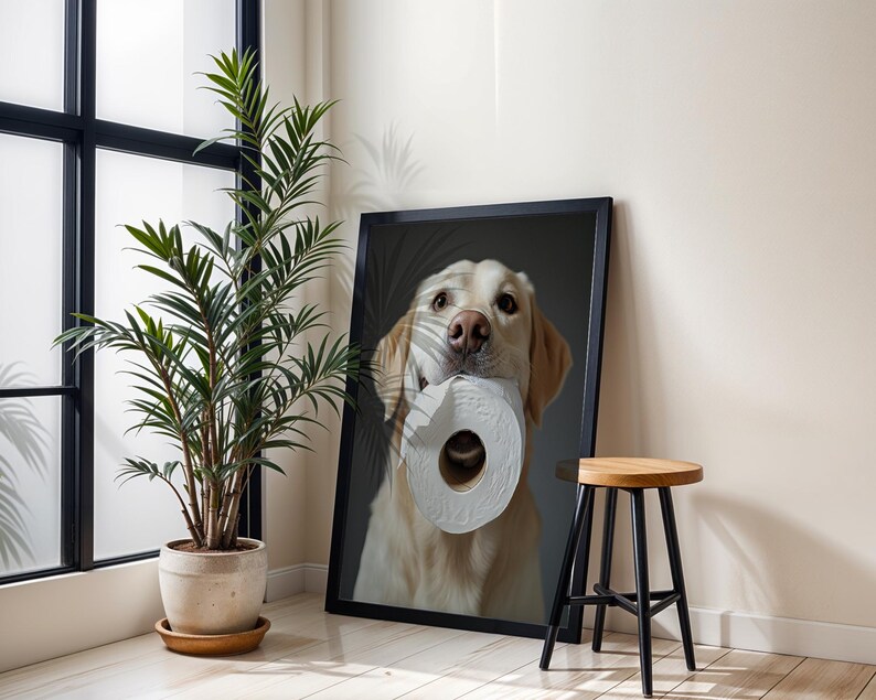 May include: A framed print featuring a golden retriever holding a roll of toilet paper in its mouth. The dog is centered in the frame against a dark background. The print is displayed next to a potted plant and a small wooden stool.