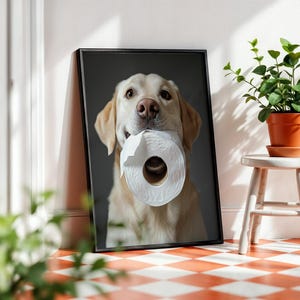 May include: Framed print featuring a golden Labrador Retriever holding a roll of white toilet paper in its mouth. The dog is centered against a dark gray background. The print is displayed against a white wall with a small plant.
