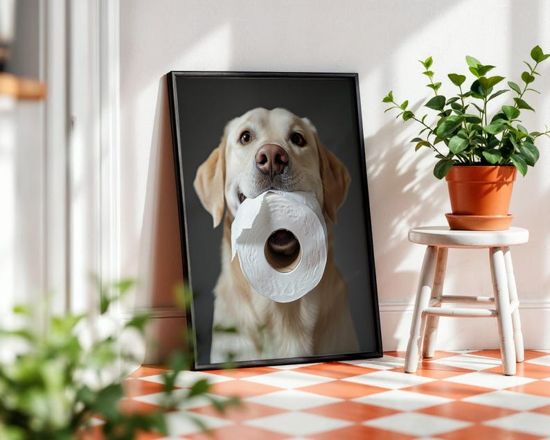 May include: Framed print featuring a golden Labrador Retriever holding a roll of white toilet paper in its mouth. The dog is centered against a dark gray background. The print is displayed against a white wall with a small plant.