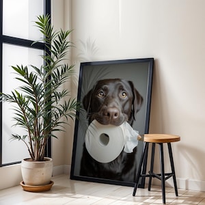 Puede incluir: Impresi&oacute;n enmarcada de un Labrador Retriever chocolate sosteniendo un rollo de papel higi&eacute;nico en la boca. El perro tiene pelaje marr&oacute;n y est&aacute; sobre un fondo neutro. Tambi&eacute;n se ven una planta en maceta y un taburete de madera.