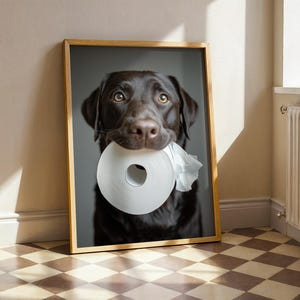 Puede incluir: Impresi&oacute;n enmarcada de un Labrador Retriever color chocolate sosteniendo un rollo de papel higi&eacute;nico blanco en la boca. El perro tiene ojos marrones y pelaje marr&oacute;n oscuro. El marco es de madera clara y el fondo gris neutro.