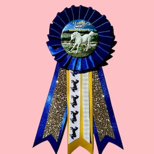 May include: A blue pleated award rosette featuring a central button with a white horse and the text I Collect BREYER Model Horses. The rosette has three long ribbons: two outer gold glitter ribbons and a central white ribbon with black horse silhouettes.