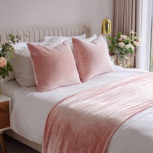 May include: A bed with white linens, two pink velvet pillows, and a matching pink velvet throw blanket. The headboard is a light beige color. A gold pendant light hangs above the bed, and a vase of flowers sits on the nightstand.