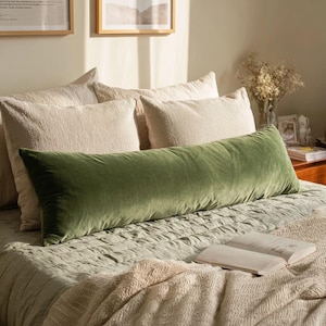 May include: A long, olive green velvet body pillow rests on a bed with several beige pillows. The bed is covered with a light green comforter and a patterned blanket. An open book is visible on the bed.