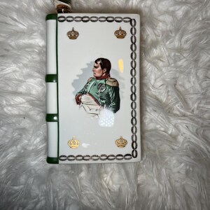 May include: A rectangular white ceramic flask with a green and gold stopper. The flask features a portrait of Napoleon Bonaparte, gold crown emblems, and a decorative border. The flask is approximately 13 cm tall.
