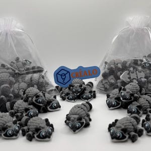 May include: A collection of gray and black raccoon figurines with blue eyes are scattered around two clear drawstring bags filled with similar figurines. A blue and orange logo with the word "CREALO" is also present.