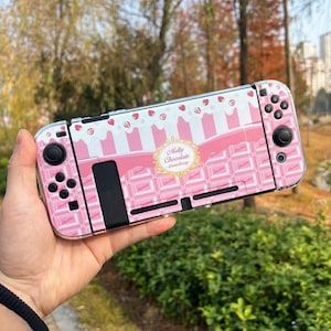 Könnte beinhalten: Eine rosa-weiße Nintendo Switch-Hülle mit Schokoladen- und Erdbeer-Design. Die Hülle hat ein sich wiederholendes Muster aus Schokoriegeln und Erdbeeren, mit den Worten "Melty Chocolate Strawberry" in einem goldenen Emblem.