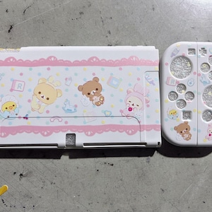 Op de afbeelding: Een witte Nintendo Switch-hoes en controller met een roze kanten rand en cartoonberen en kuikens. De controller heeft een glitterontwerp met pootafdrukken en cirkels.