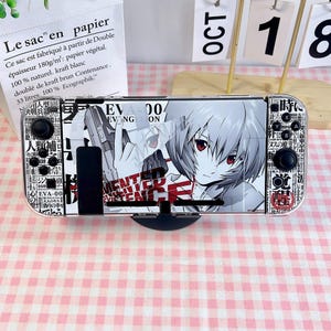 Può includere: Una custodia protettiva trasparente per console di gioco portatile, con grafica anime in bianco e nero con accenti rossi. La custodia presenta testo giapponese e le parole "EVA-00" e "MENTED SILENCE". La console è su un supporto nero.