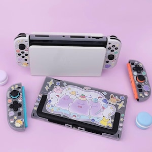 Może przedstawiać: Konsola Nintendo Switch i akcesoria, w tym stacja dokująca, kontrolery i etui ochronne. Kontrolery i etui mają kolorowe wzory postaci z kreskówek. Tło jest jednolite, jasnofioletowe.