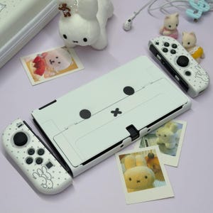 May include: A white gaming console with a minimalist rabbit face design on the back, accompanied by two matching white controllers featuring black buttons, small rabbit graphics, and star patterns, surrounded by small plush rabbit figurines.