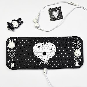 Op de afbeelding: Een zwarte gameconsole-hoes met witte stippen en een centrale hartvormige afbeelding met drie Miffy-figuren en de tekst Miffy. De set bevat witte bedrade oordopjes met sterpatronen, een gepixelde karakterspeld en witte duimgrepen voor de controllers.