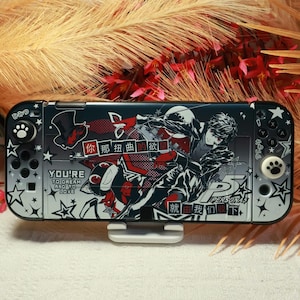 May include: A black and silver Nintendo Switch console with a custom design featuring anime-style artwork and text. The design includes a character illustration, the text "PERSONA 5", and other Japanese text. The console has paw-shaped buttons.