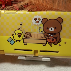 Puede incluir: Funda para Nintendo Switch a cuadros amarillos y blancos con personajes de dibujos animados. Un oso pardo y un pollito amarillo juegan a un juego de mesa. La funda tiene un cierre blanco y está decorada con texto japonés.