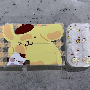 Pode incluir: Uma capa estampada em amarelo e castanho com um personagem de cachorro de desenho animado com boina e um personagem de cachorro menor. Ao lado, uma capa transparente decorada com desenhos animados de comida e personagens.