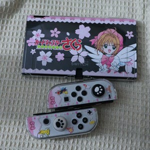 Op de afbeelding: Een Nintendo Switch-console en Joy-Con-controllers met een aangepast ontwerp. De consolehoes is zwart met roze accenten, kersenbloesemafbeeldingen en Japanse tekst. De Joy-Cons zijn lichtroze met karakterillustraties.