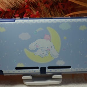 Op de afbeelding: Een lichtblauwe Nintendo Switch-hoes met een stripfiguur die op een halve maan slaapt. De hoes heeft een ontwerp met een hemelthema met wolken, sterren en een kleine roze beer. De hoes is ontworpen om de Switch-console te beschermen.