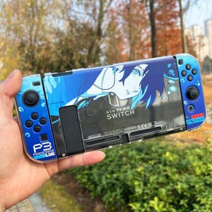 May include: A Nintendo Switch console with a custom blue and white Persona 3 Reload design. The design features an anime character and the text "NINTENDO SWITCH" and "P3 PERSONA 3 RELOAD". The controllers are blue.