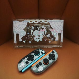 May include: Clear plastic Nintendo Switch case and two Joy-Con controllers. The case has a pink curtain design with cartoon characters and musical notes. The Joy-Cons are grey with pink and white character stickers.
