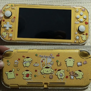 May include: Yellow handheld games console with a clear protective case. The case and console feature a repeating pattern of cartoon characters, desserts, and stars. The screen is black. The text "Pom Pom Purin" is visible.
