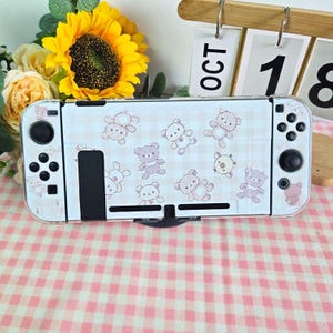 May include: A white Nintendo Switch console with a decorative skin featuring a pattern of light pink teddy bears on a light blue and white checkered background. The console is set against a backdrop of flowers and a calendar displaying "OCT 18".