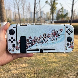 Könnte beinhalten: Weiße Handheld-Spielkonsole mit farbenfrohem Grafikdesign mit Zeichentrickfiguren. Die Konsole hat schwarze Steuerknüppel und Tasten. Die Grafik enthält den Text "Pocket Monster".